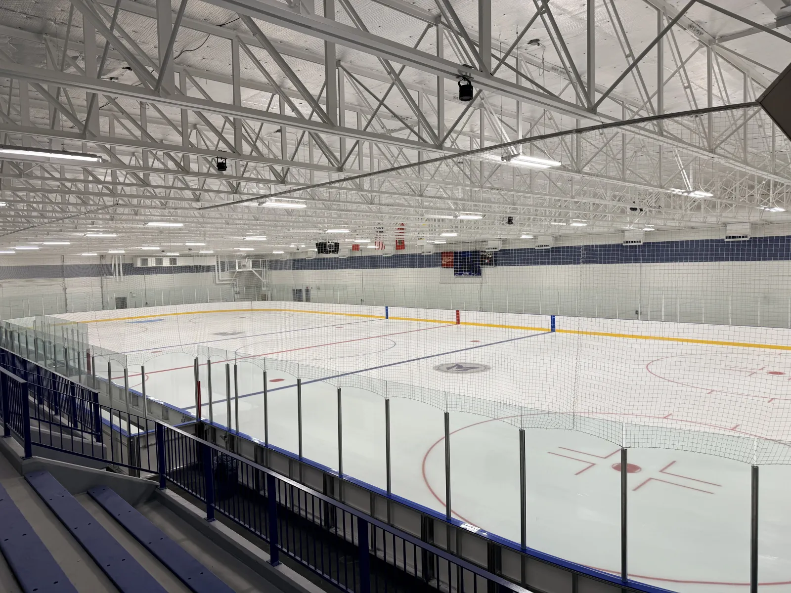 Empty ice sheet viewed from the upper seating area, showing the full rink with glass, netting, and a scoreboard under steel roof trusses. — Brandon Community Sportsplex