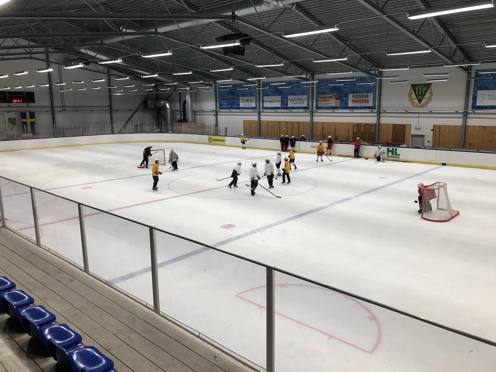 The whole ice sheet at Brantbrinks ishall in Tullinge seen from the stands, with blue and red lines, blue sponsor boards, steel roof trusses and a small group skating at the centre (listing name Ica Riksten Arena, same coordinates) — Brantbrinks ishall (Tullinge)