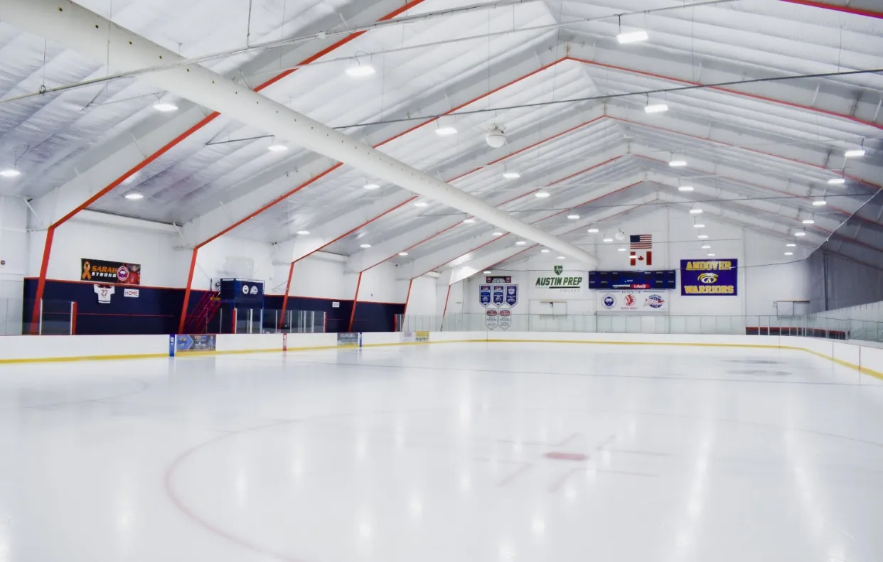 An empty rink under a white insulated roof with red trusses, a "Sarah Strong" banner on the left wall and team banners and flags at the far end. — Breakaway Ice Center