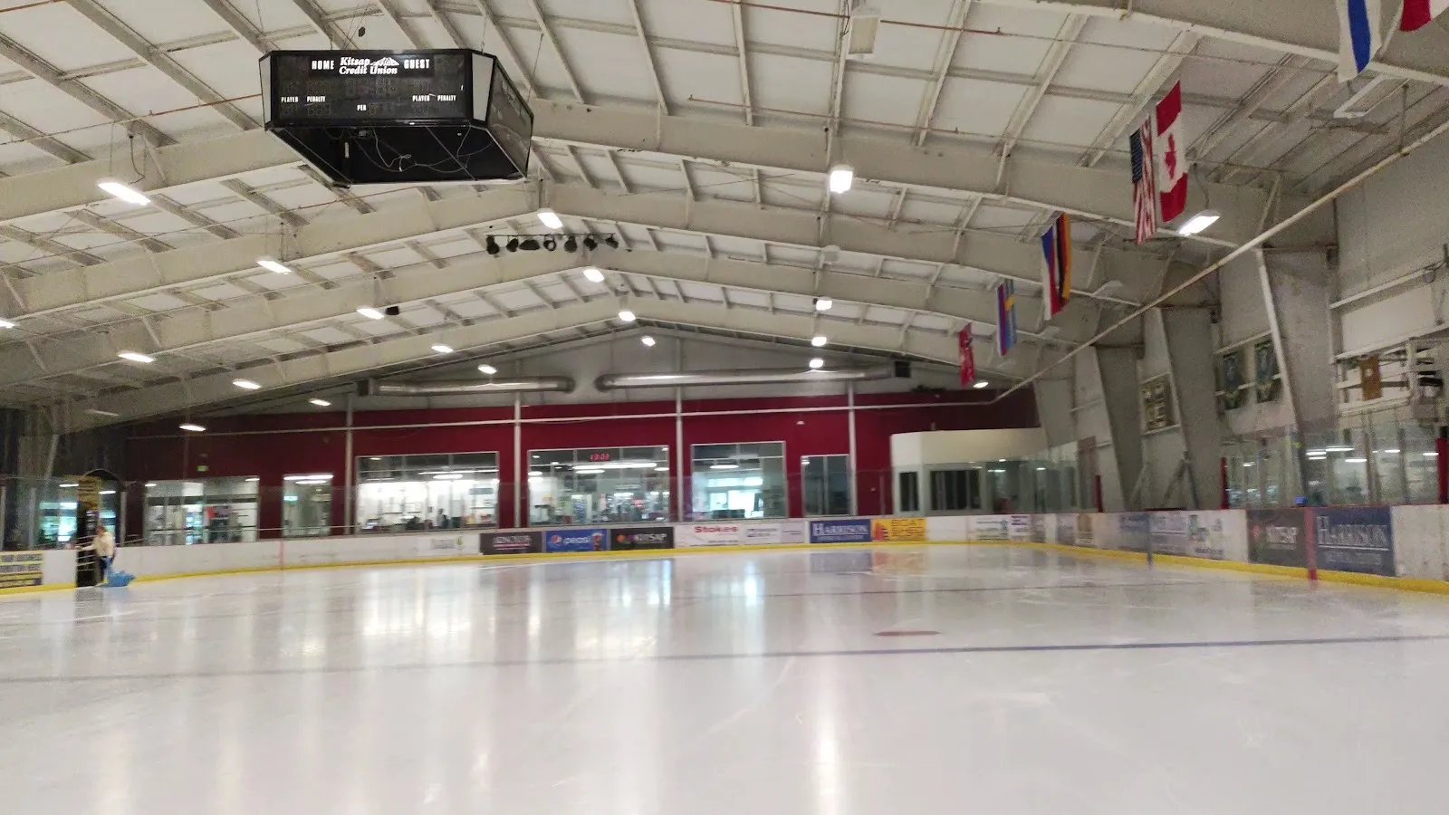 The empty Bremerton Ice Center sheet under white roof trusses, with the hanging scoreboard, a row of flags and the red-walled lobby windows — Bremerton Ice Center