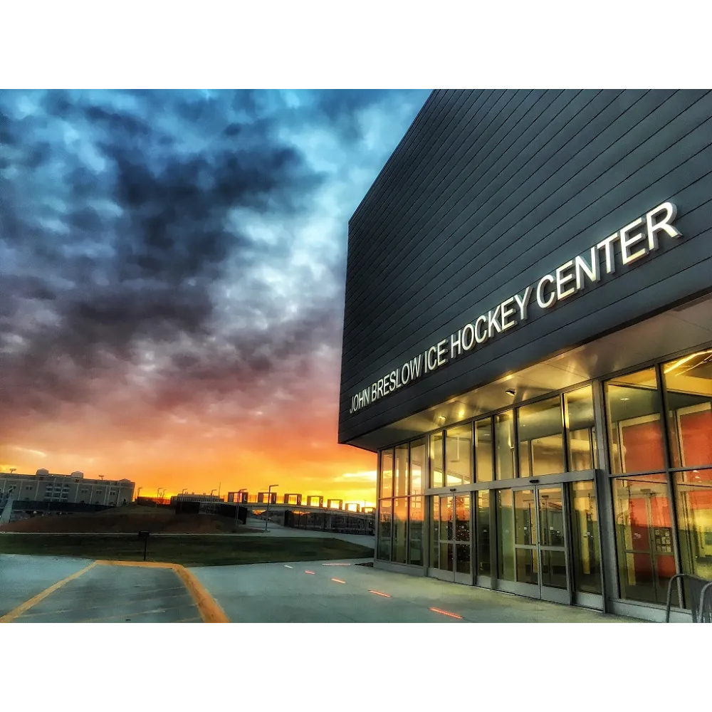 Front entrance of the building at sunset, with an illuminated 'JOHN BRESLOW ICE HOCKEY CENTER' sign on the dark metal-clad facade and glass doors below. — John Breslow Ice Hockey Center