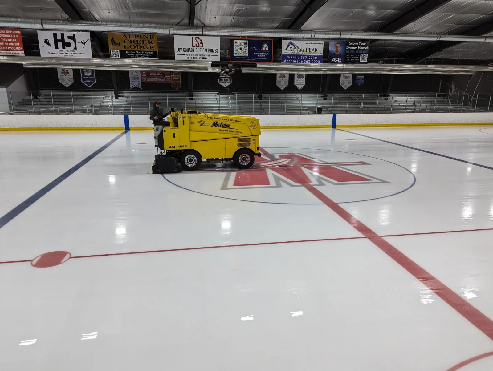 Full ice sheet inside The Brett, an ice resurfacer driven by staff parked at center ice over the red 'M' logo, bleacher seating and sponsor banners along the boards, 'THE BRETT' sign visible above the stands. — Brett Memorial Ice Arena