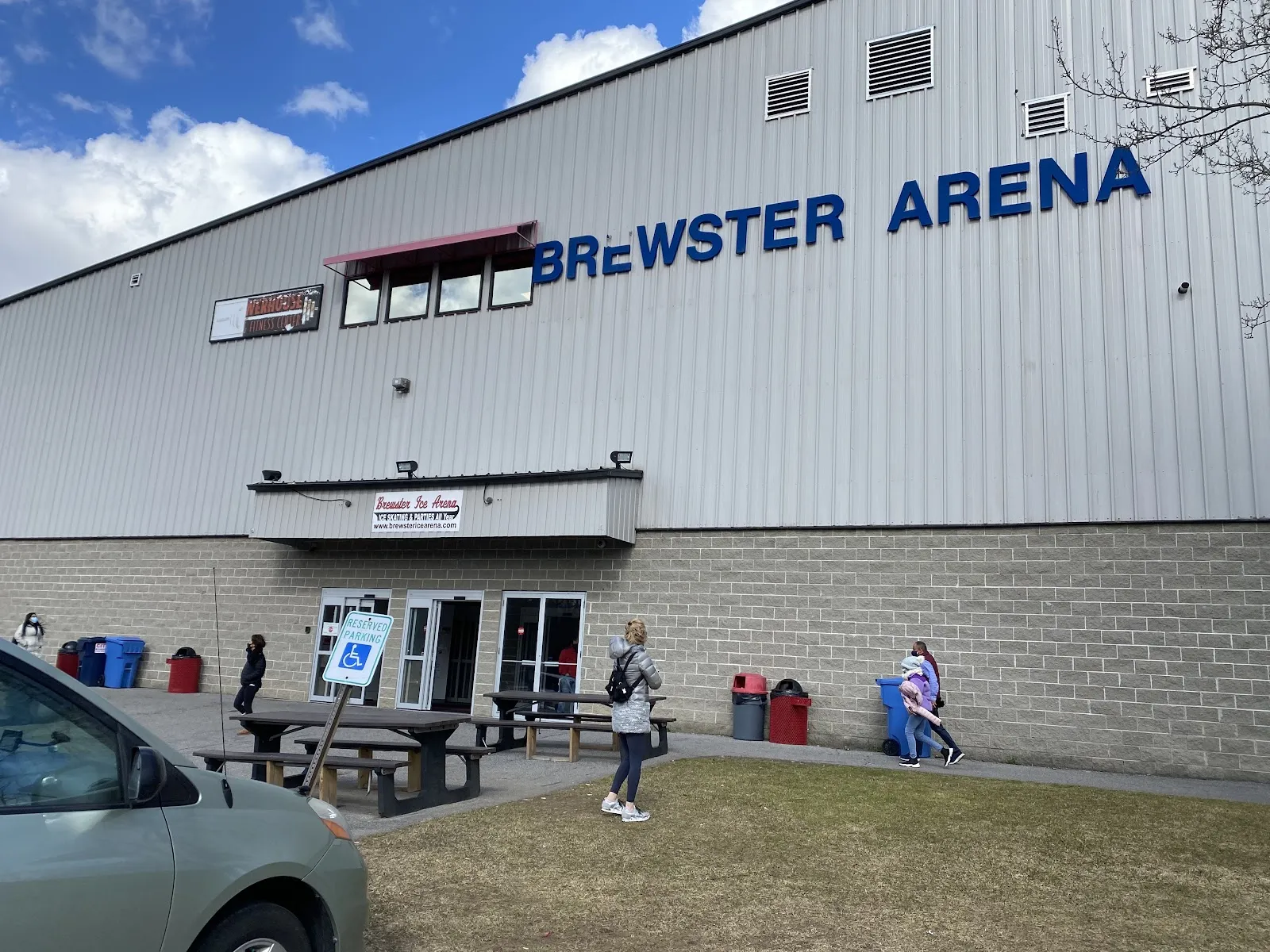El muro de metal gris de Brewster Arena con su nombre en letras azules y la entrada debajo, visto desde el césped — Brewster Ice Arena