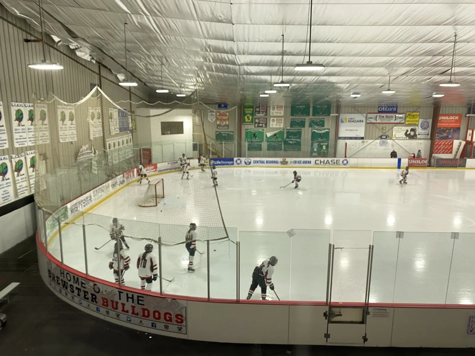 Un partido visto desde las gradas, con jugadores repartidos por la pista dentro de un barandal rojo curvo y banderines de los equipos colgados en la pared ondulada del fondo. — Brewster Ice Arena