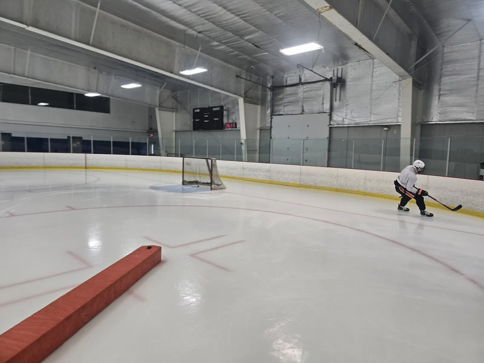A wide view of the ice from the end boards, a red-edged bench in the foreground and a lone player at the far side under a steel-framed ceiling. — BIGGBY COFFEE Ice Cube - Brighton