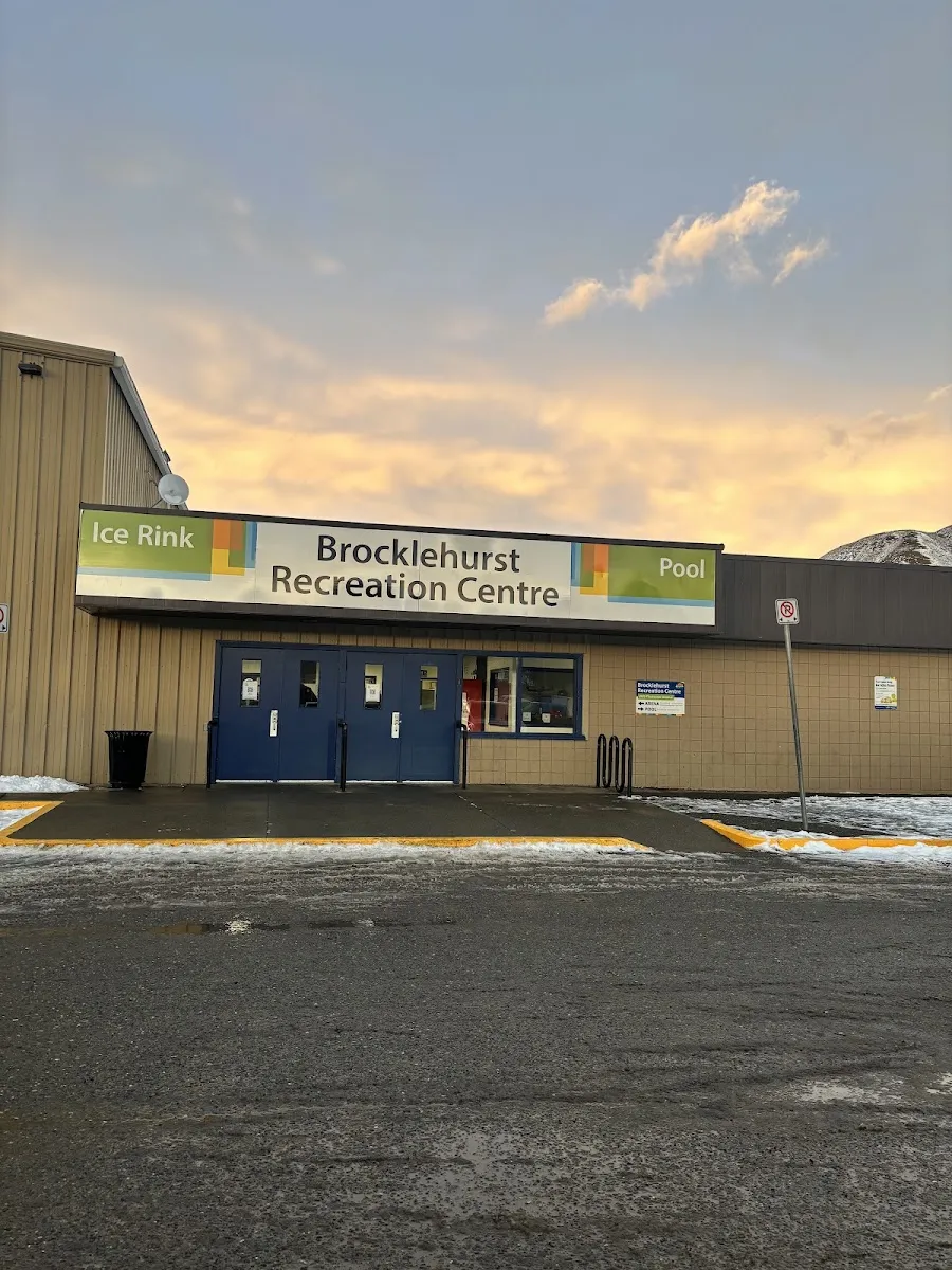 La fachada revestida en tono arena del Brocklehurst Recreation Centre, con su rótulo de Ice Rink y Pool sobre dobles puertas azules, nieve junto al bordillo y un cielo de atardecer rosa y dorado detrás. — Brocklehurst Arena