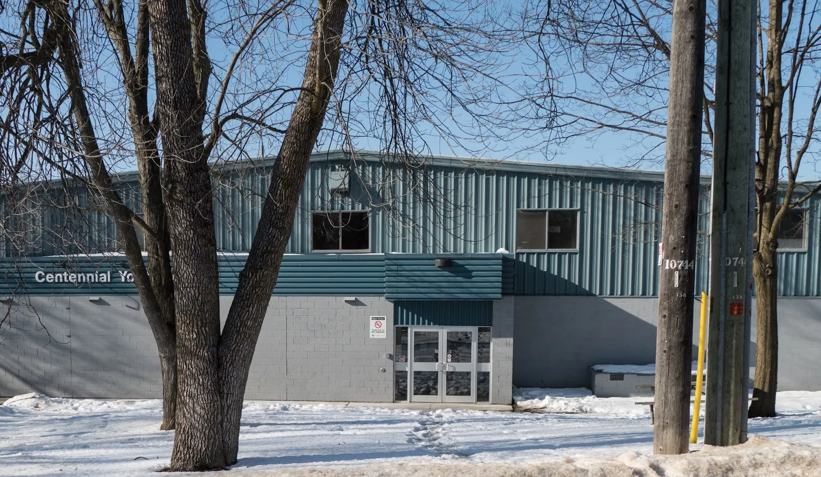 El frente verde azulado con revestimiento metálico de Brockville Centennial Youth Arena en invierno, con sus puertas de entrada y el nombre de la arena a lo largo del muro detrás de árboles sin hojas — Brockville Centennial Youth Arena