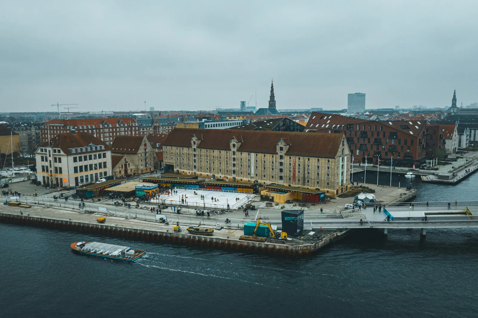 The small white rink on the harbour quay in Copenhagen seen from above, with the brick warehouse buildings of Christianshavn and the water around it. — Broens Skøjtebane