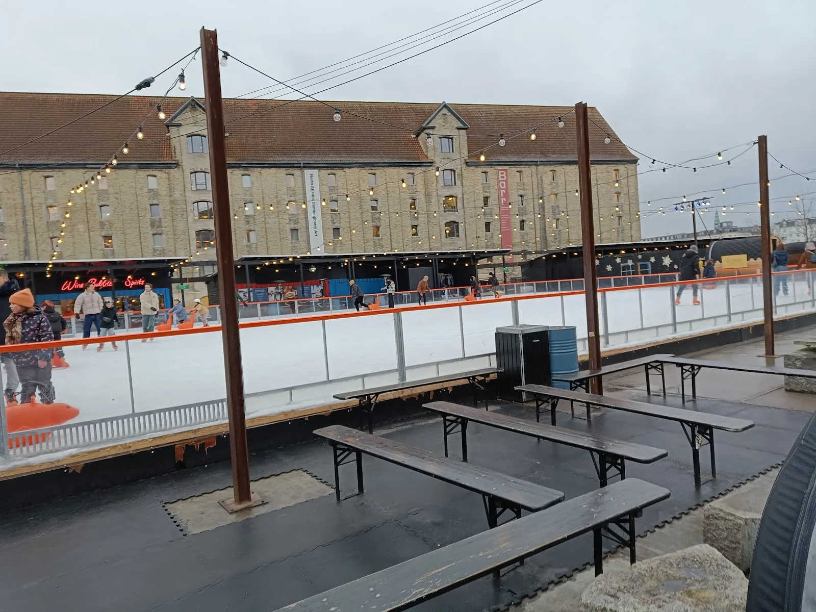 The rink's boards and ice seen from the benches on the quay, with strings of lights over the rink and the old brick warehouse behind. — Broens Skøjtebane