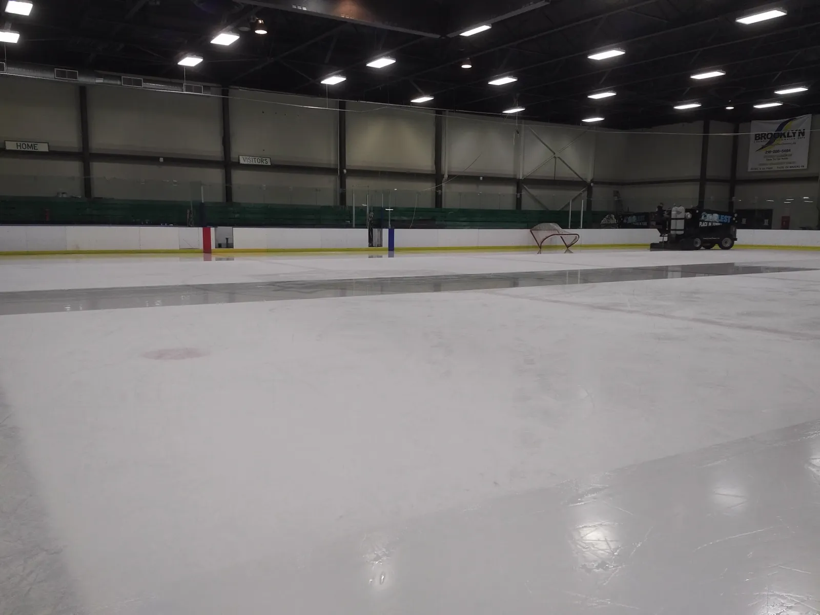 La pista vacía bajo un techo oscuro con hileras de luces, con las vallas y el cristal del fondo y una pulidora de hielo estacionada en el extremo opuesto. — John M. Coyne Recreation Center (Brooklyn)