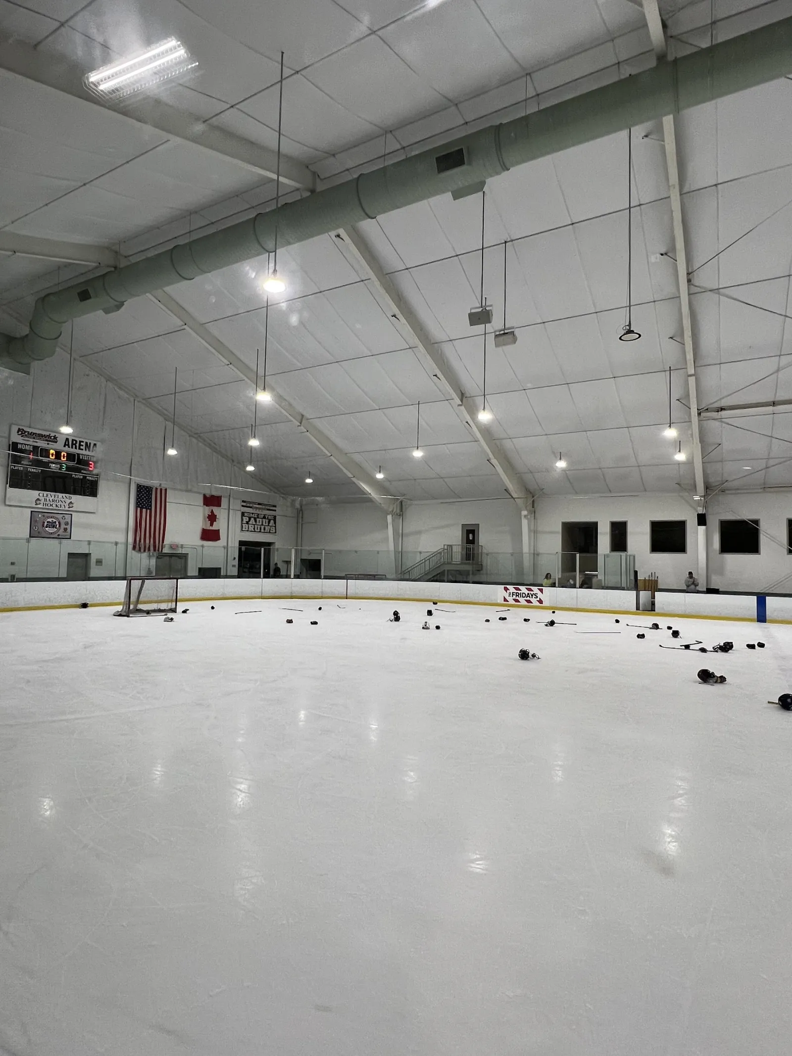 The empty ice after a practice with pucks scattered on it, American and Canadian flags, a Walsh Jesuit hockey banner and the scoreboard on the far wall. — Brunswick Auto Mart Arena (Strongsville)