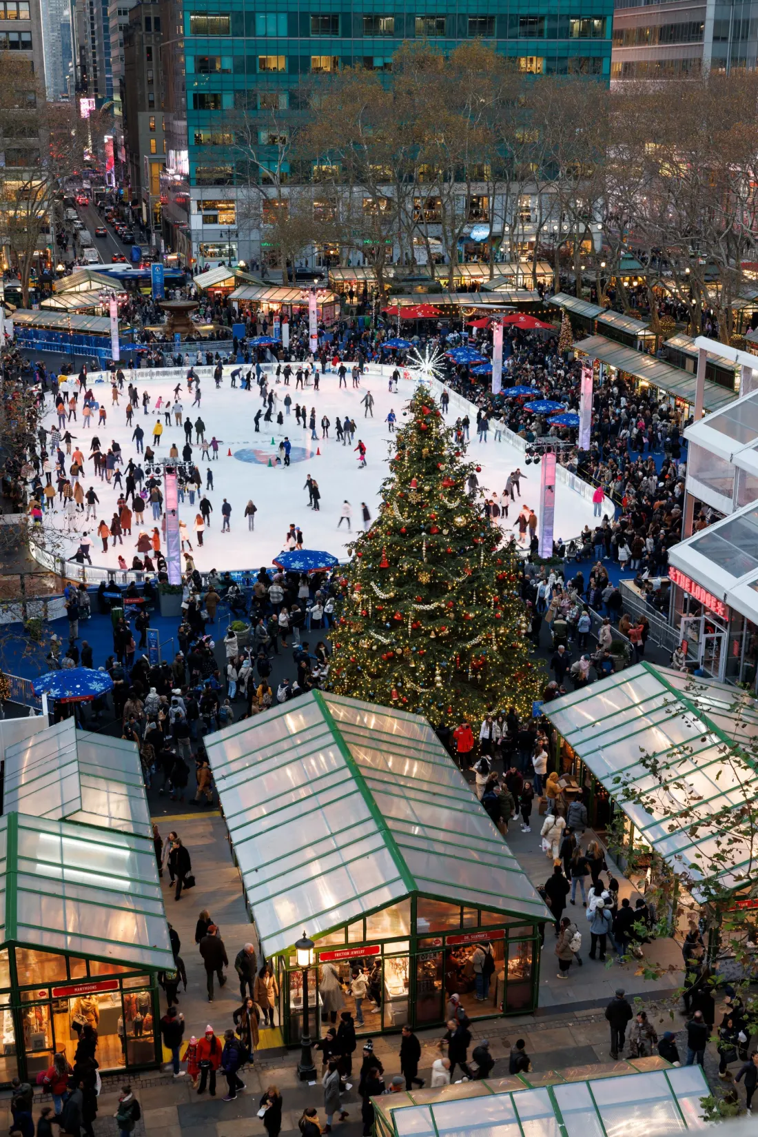 Winter Village from above: the rink on the park lawn with the tall Christmas tree, the glass-roofed holiday shop kiosks in the foreground and Midtown towers around the park. — The Rink at Bryant Park