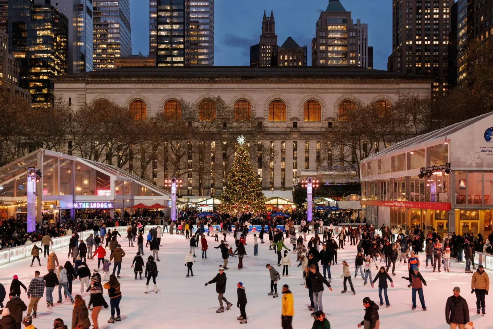 The rink full of skaters at dusk, with The Lodge's glass pavilion, the Christmas tree and the New York Public Library's facade behind. — The Rink at Bryant Park