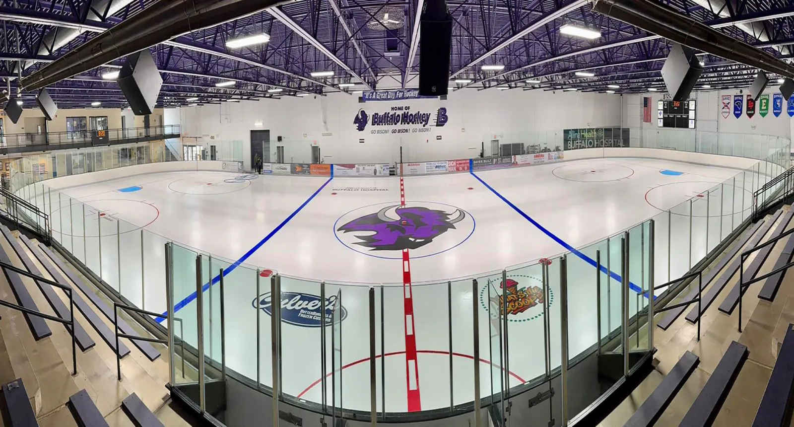 A wide view of the empty main rink from the stands, with the Bison logo at center ice and Buffalo Hockey banners on the far wall. — Buffalo Civic Center