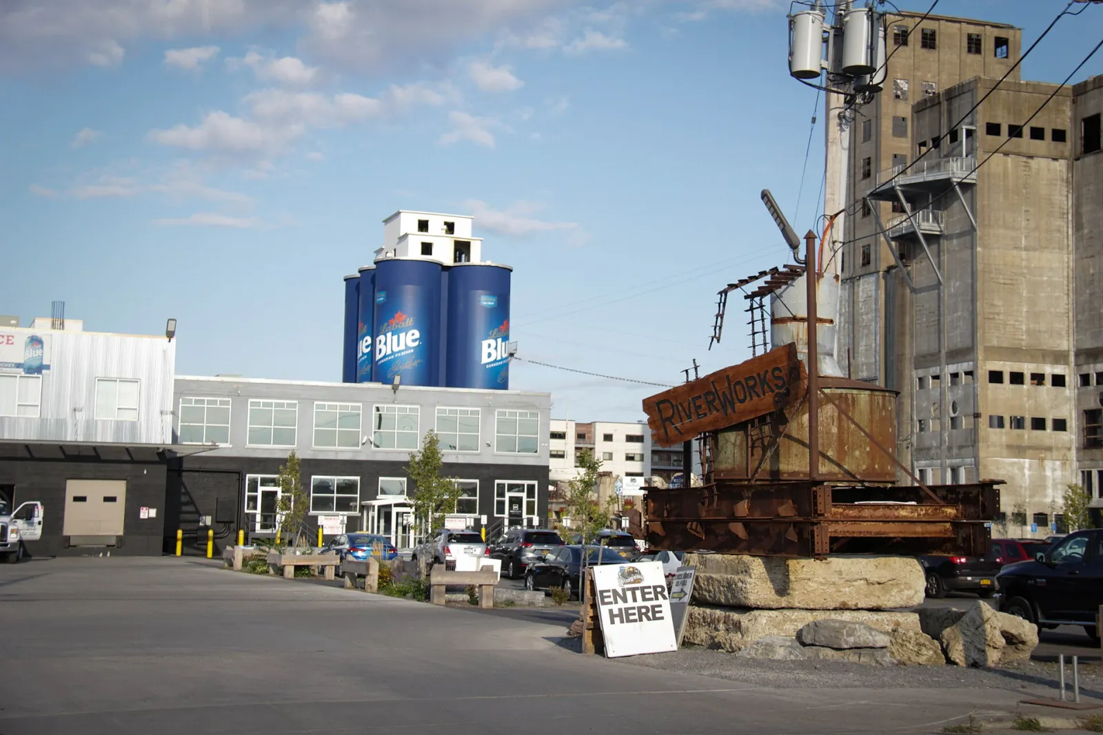 La entrada de RiverWorks con su letrero oxidado de RiverWorks sobre una base de roca y un tablero ENTER HERE, frente a silos de granos pintados como latas de Labatt Blue. — Buffalo RiverWorks