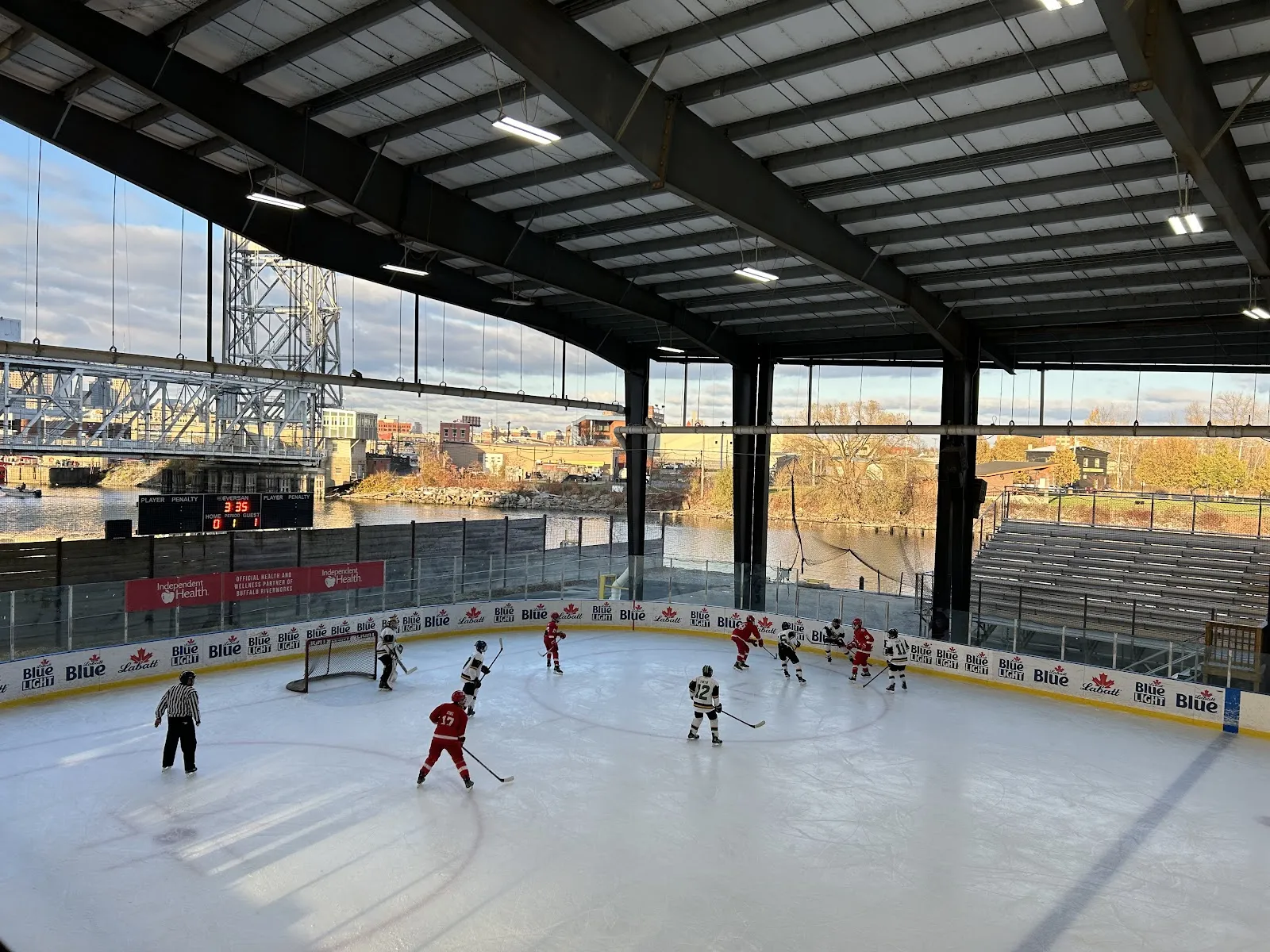 La pista de hielo cubierta bajo un techo con estructura de acero abierto hacia el lado del río, con un grupo de patinadores jugando un partido de hockey sobre el hielo y el puente levadizo y los elevadores de granos visibles al otro lado del vidrio. — Buffalo RiverWorks