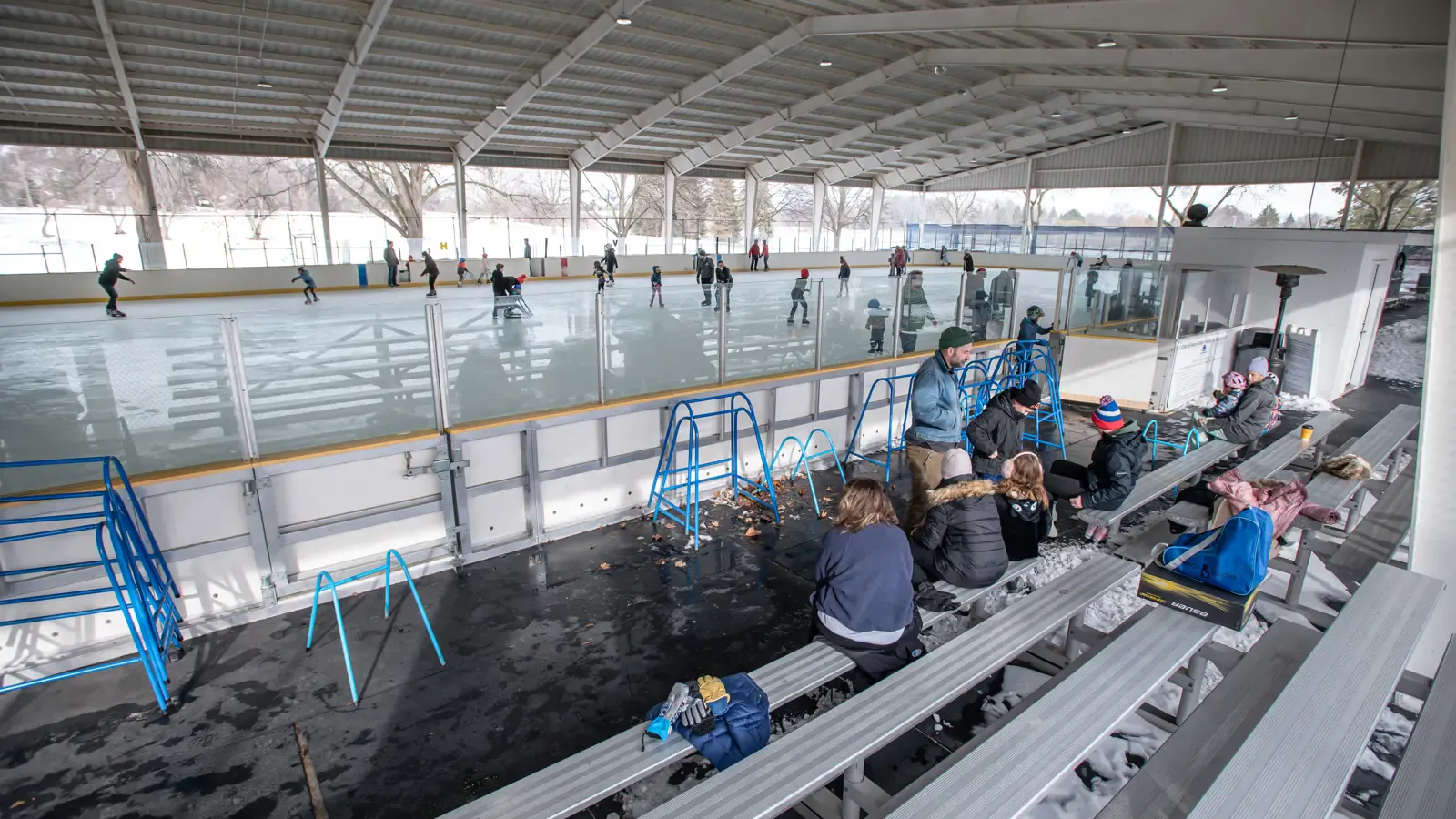 Skaters on the covered Buhr Park ice rink seen from the bleachers, where families sit under a steel roof along the boards. — Buhr Park Outdoor Ice Arena