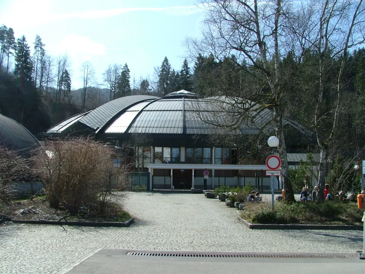 Das dunkle, gewölbte Dach der Eishalle, über einen gepflasterten Vorplatz gesehen, dahinter kahle Birken und bewaldete Hügel. – Bundesstützpunkt Füssen
