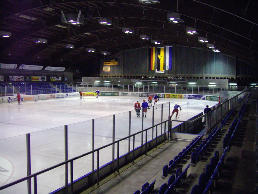 Läuferinnen und Läufer in der Arena unter dem gewölbten Dach, an der Seite blaue Tribünensitze und im Vordergrund die Bande. – Bundesstützpunkt Füssen