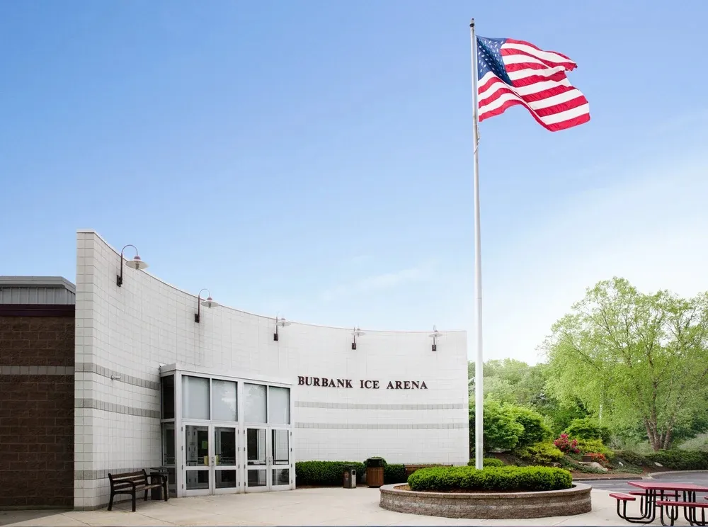 The curved white entrance wall of the Burbank Ice Arena with its name across the front, glass doors below and an American flag on a tall pole. — Burbank Ice Arena