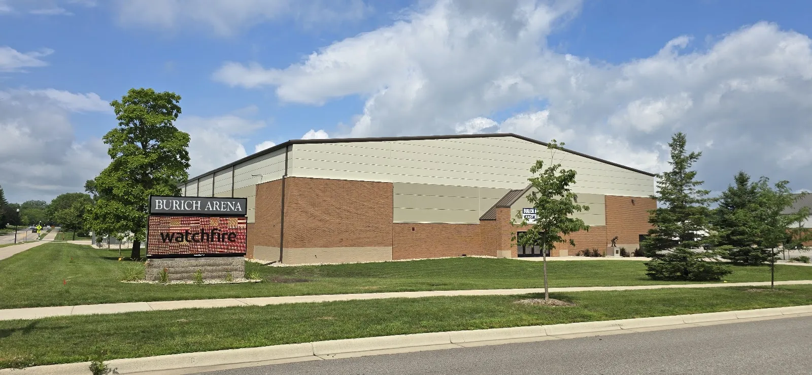 The arena from the street: a long pale-panelled and brick building with a pitched roof, and a roadside sign reading Burich Arena in front of it among small trees and lawn. — Burich Arena