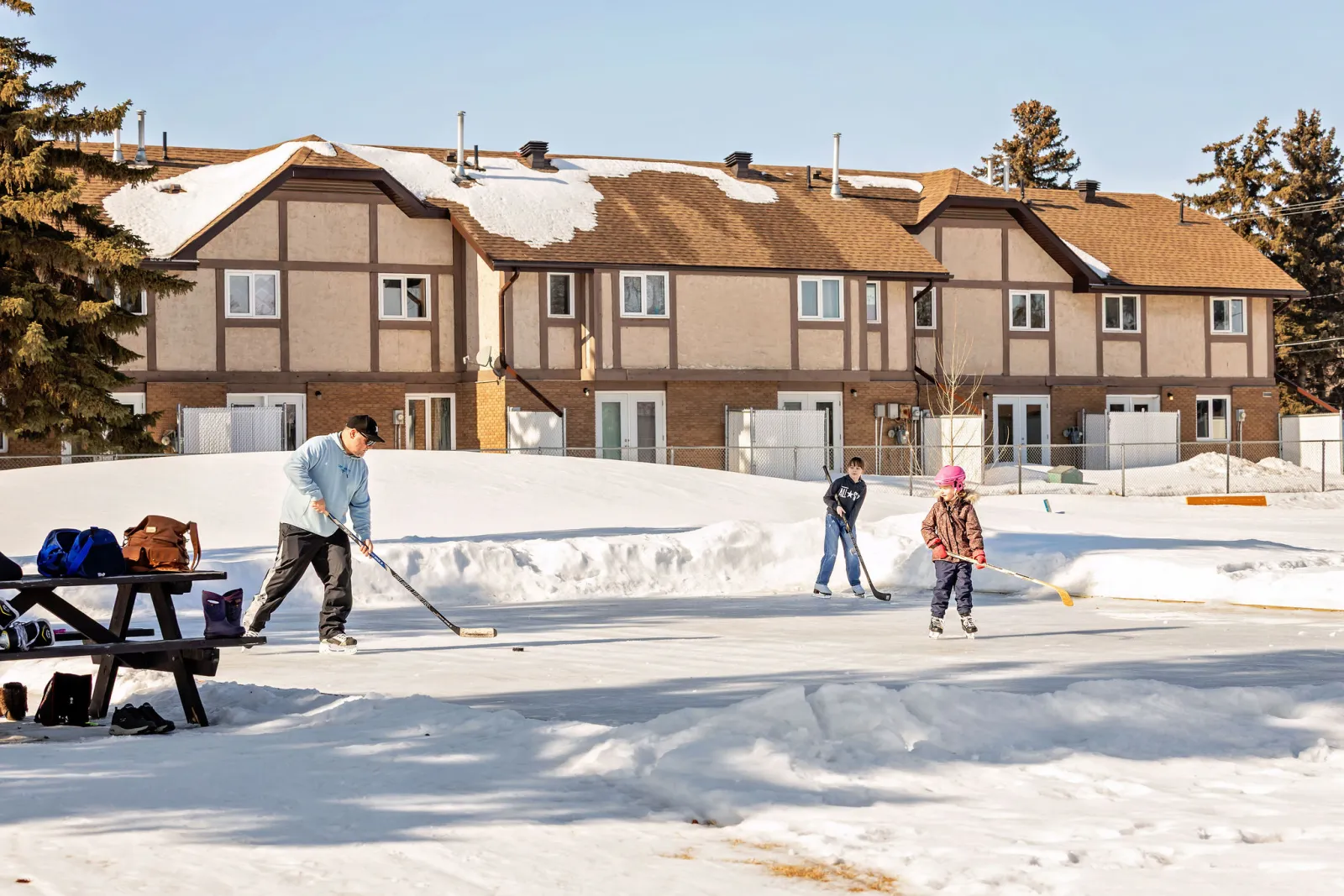 A casual game on the unboarded outdoor ice surface beside a picnic table, with snowbanks around it and townhouses behind. — Burke Playground Outdoor Ice Surface (Melfort)