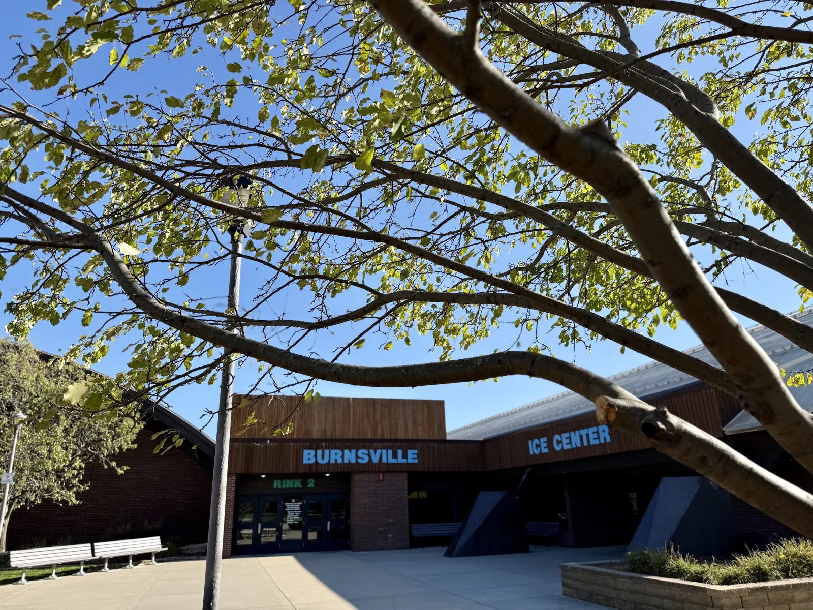 La entrada baja color café con Burnsville e Ice Center rotulados en azul sobre las puertas, enmarcada por ramas de árboles en un día soleado. — Burnsville Ice Center