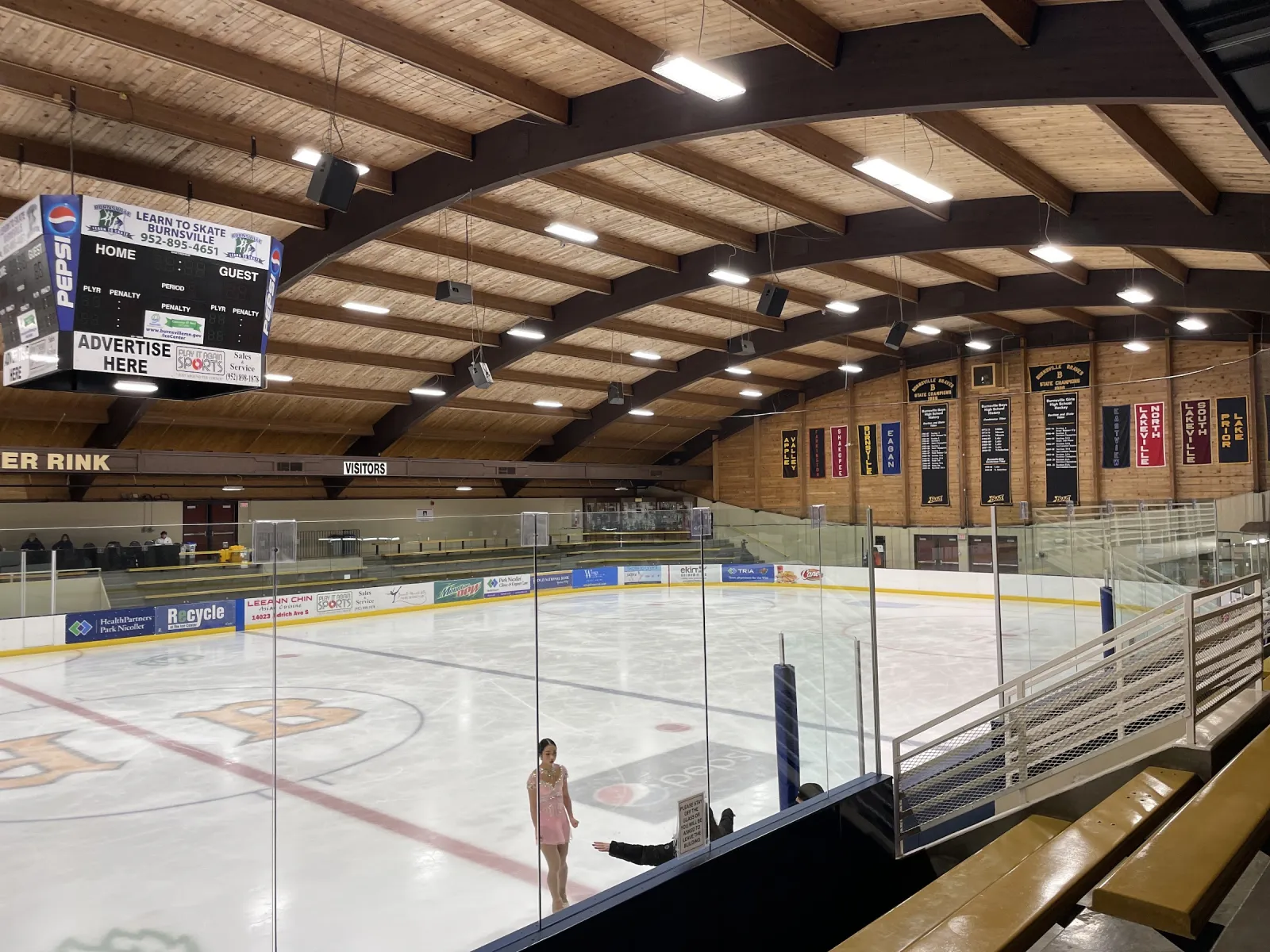 El hielo visto desde las gradas bajo un techo curvo de madera, con mantas, un marcador y anuncios de patrocinadores alrededor de la pista. — Burnsville Ice Center