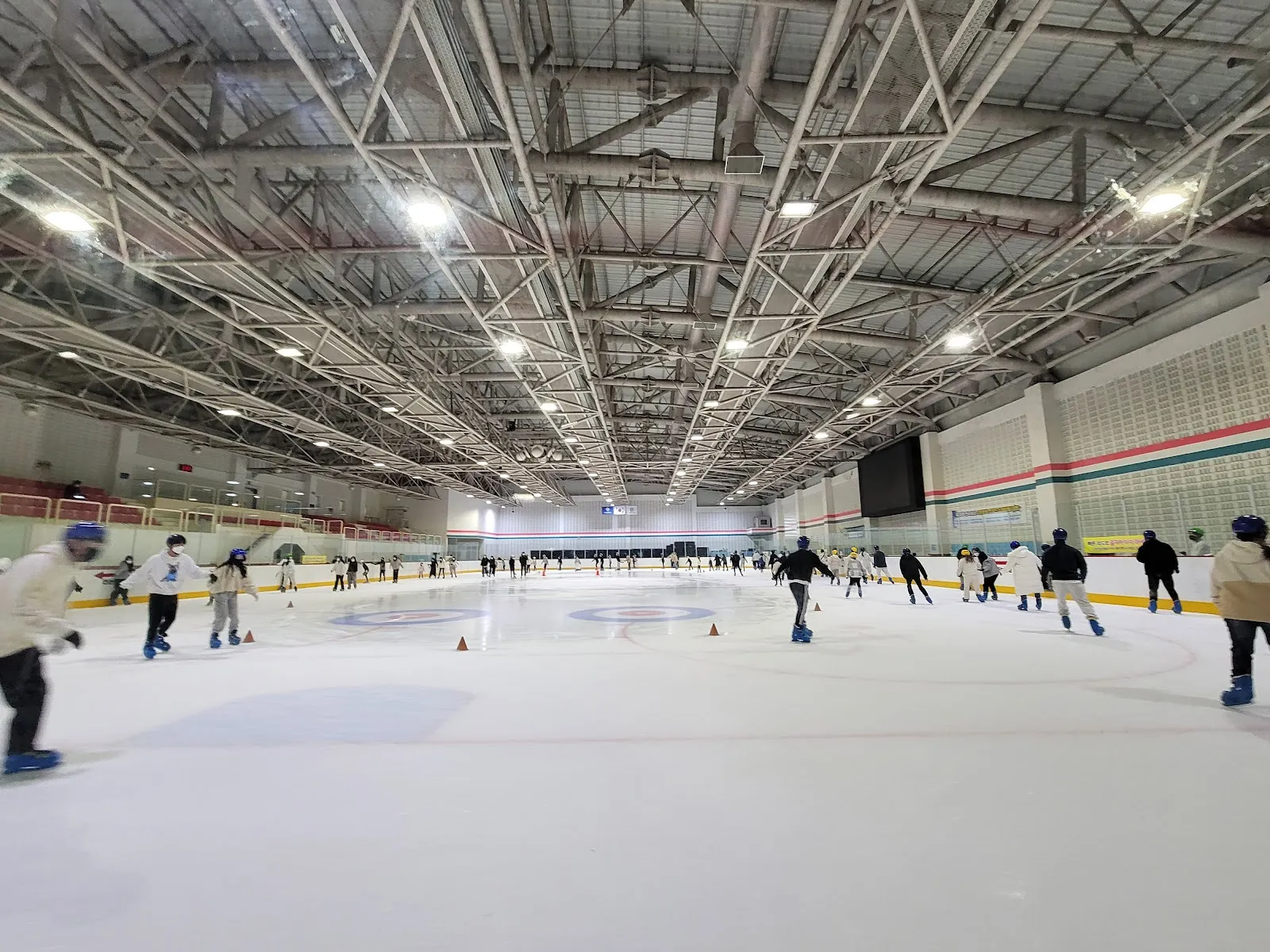 The Busan Indoor Ice Rink ice seen from the end of the sheet under the steel roof, with skaters in the distance along the boards. — Busan Indoor Ice Rink
