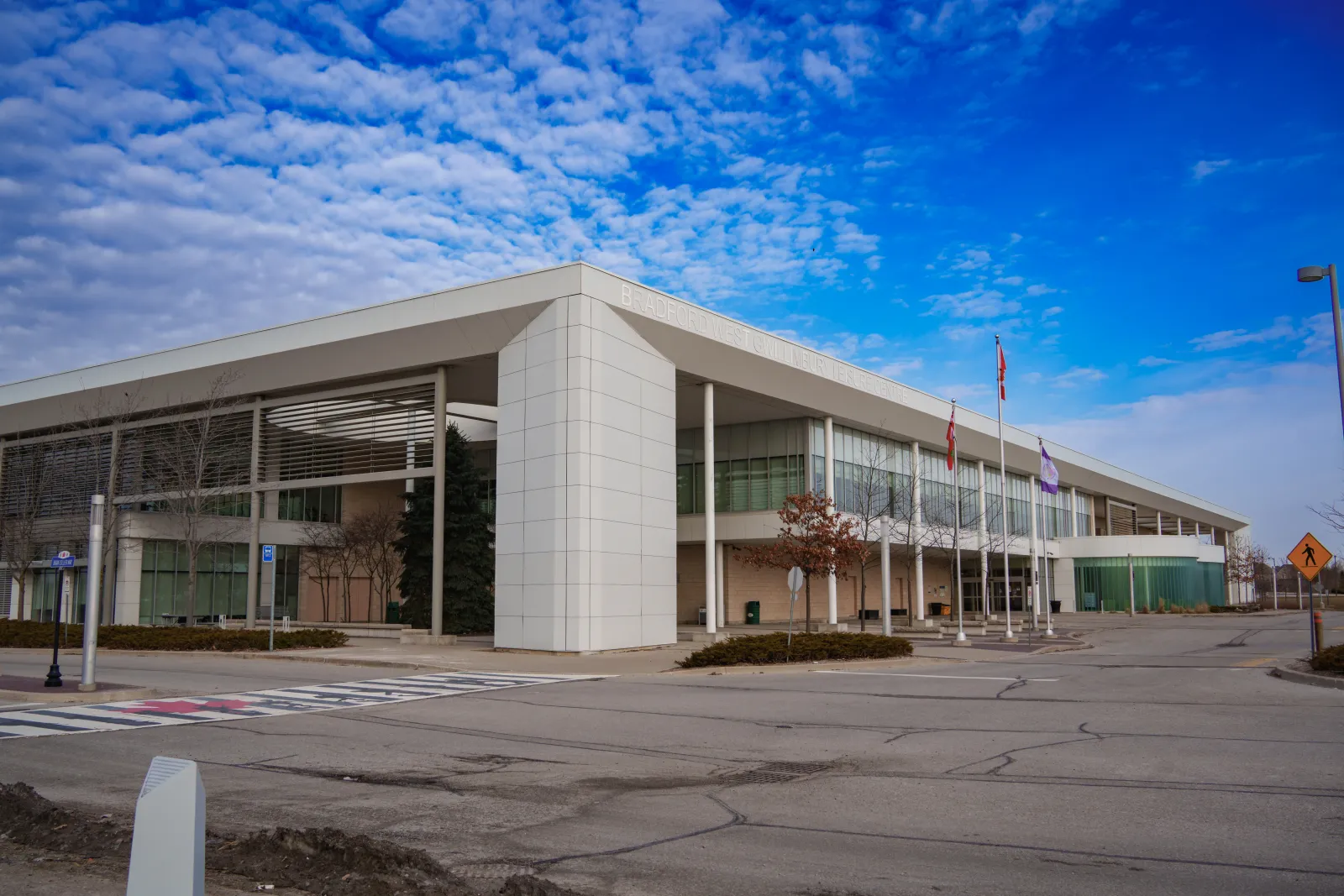 El BWG Leisure Centre, un largo edificio blanco con las letras Bradford West Gwillimbury a lo largo del borde del techo, un pilar de azulejos y tres astas de bandera. — BWG Leisure Centre (Bradford)
