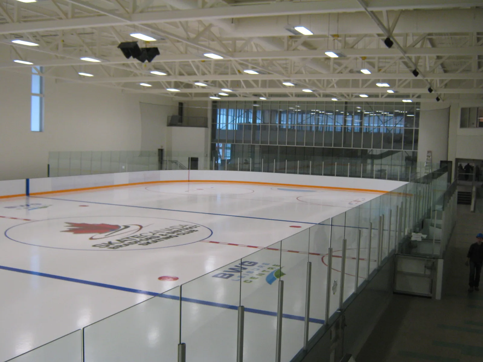 Una pista vacía desde la esquina superior: el logotipo del club de patinaje en el centro del hielo, vidrio a lo largo de las vallas y el techo blanco de armaduras de acero. — BWG Leisure Centre (Bradford)