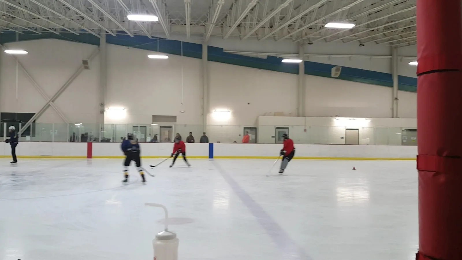 Una vista amplia de la pista desde el extremo, con unos cuantos jugadores repartidos por el hielo bajo un alto techo de acero con un poste rojo en el borde derecho. — Cabin John Ice Rink