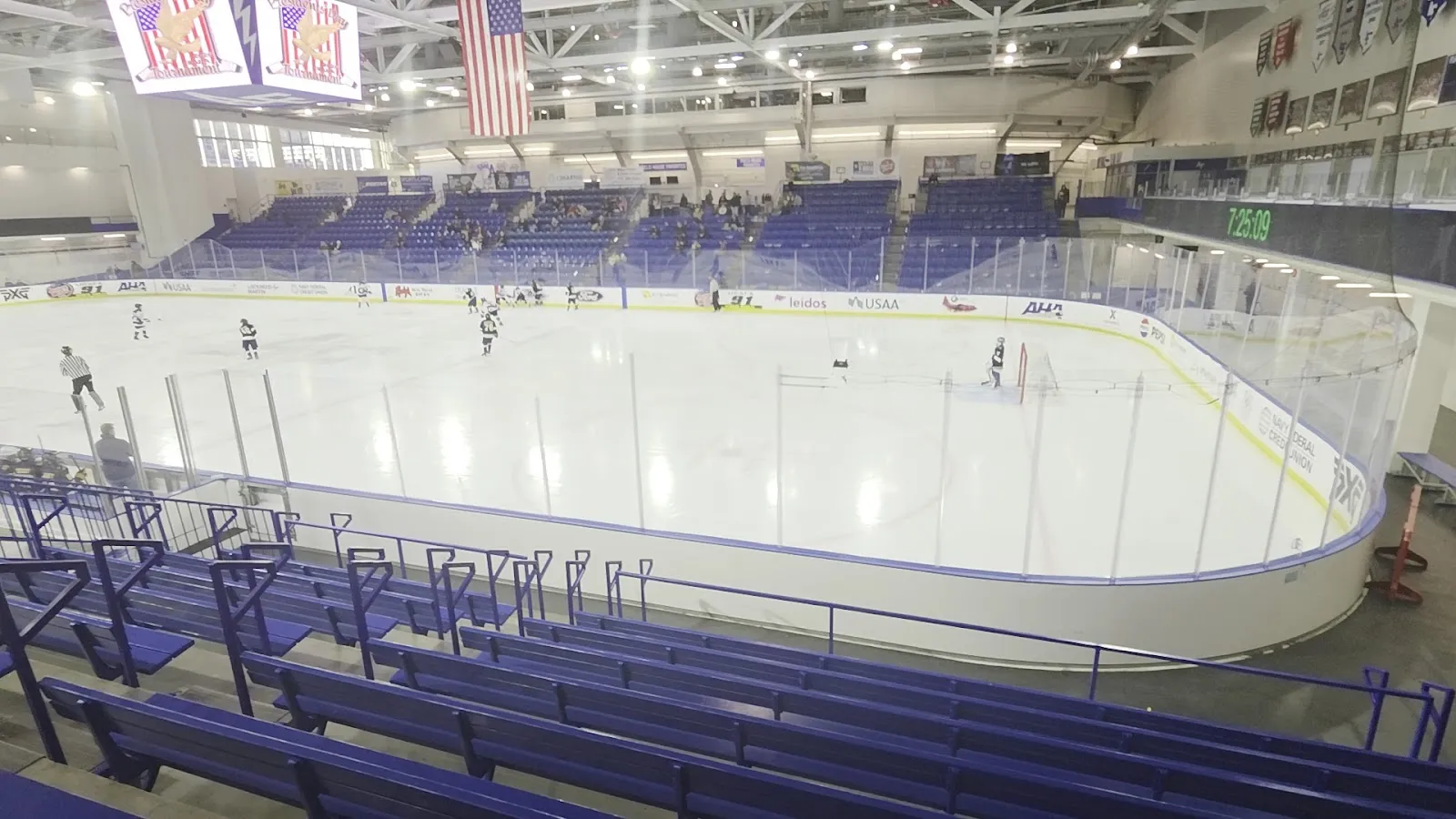 The sheet from the stands, with the American flag hanging from the roof trusses, the blue seating bowl along the far side and a few players warming up on the ice. — Cadet Ice Arena (Air Force Academy)
