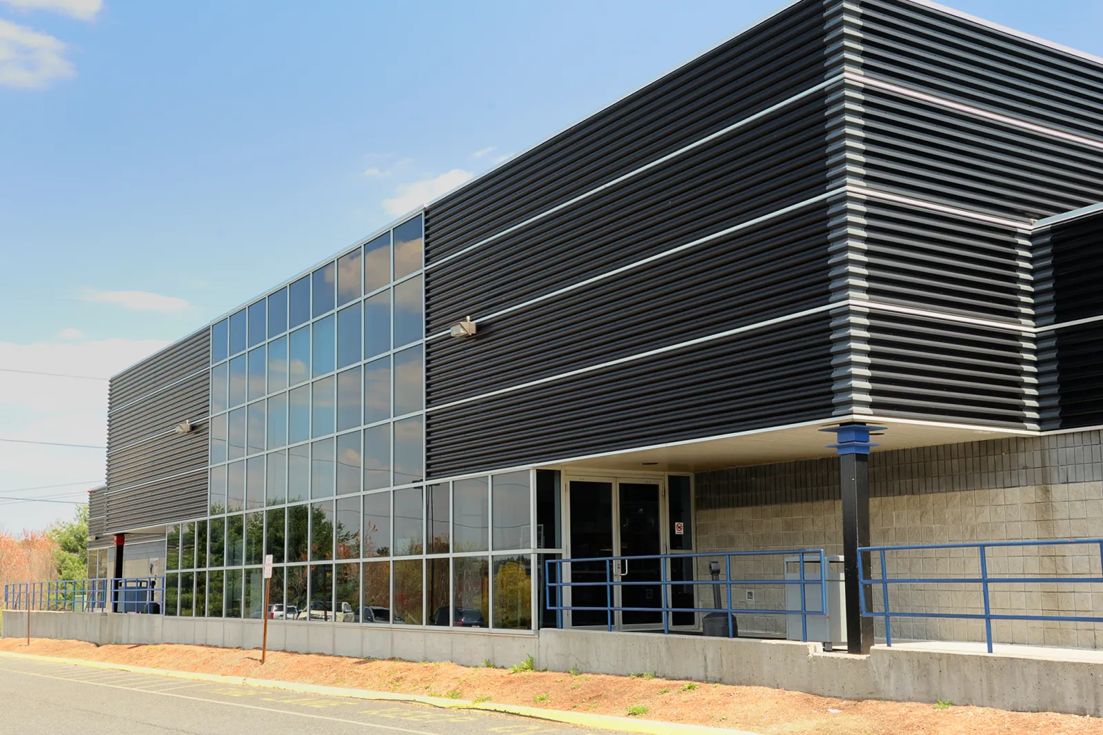 El costado de la arena: un largo edificio revestido de metal oscuro con un bloque de entrada de muro cortina de vidrio, visto desde el césped junto a la carretera. — Cadillac Ice Arena