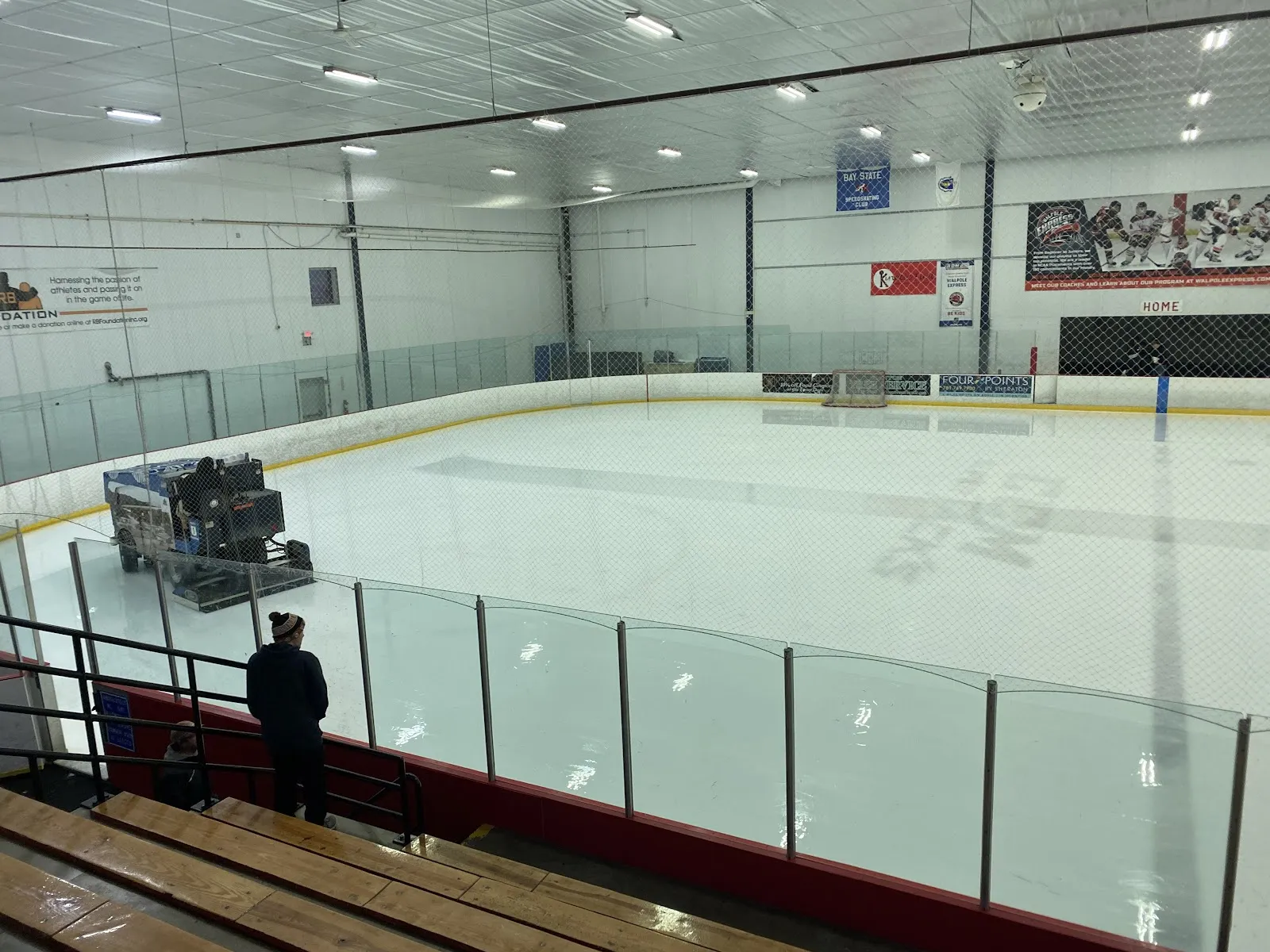 Una Zamboni renovando la superficie de la pista vacía, vista desde las gradas detrás del vidrio, con mantas a lo largo de la pared del fondo. — Cadillac Ice Arena