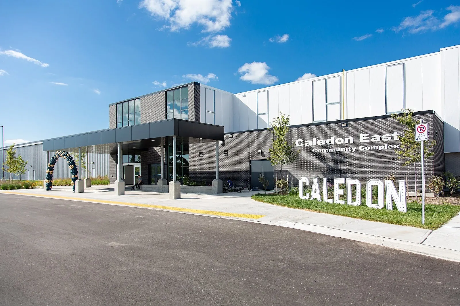 The Caledon East Community Complex entrance canopy beside a dark brick wall lettered with its name, white CALEDON letters on the lawn in front. — Caledon East Community Complex
