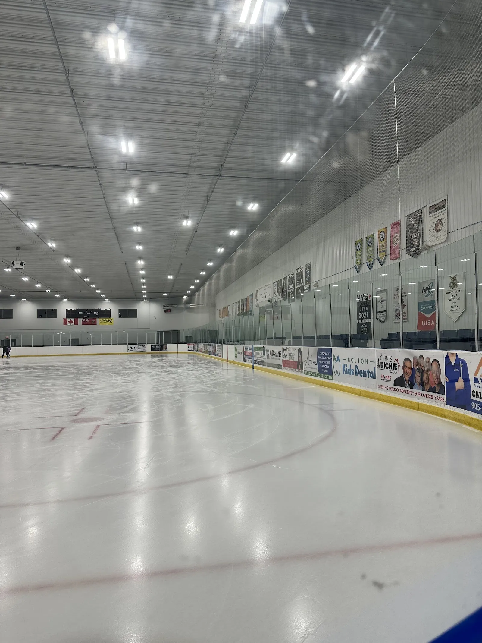 A portrait view of the empty sheet through the glass, white ice with a faint line and a ceiling lit by long fixtures above the boards. — Caledon East Community Complex