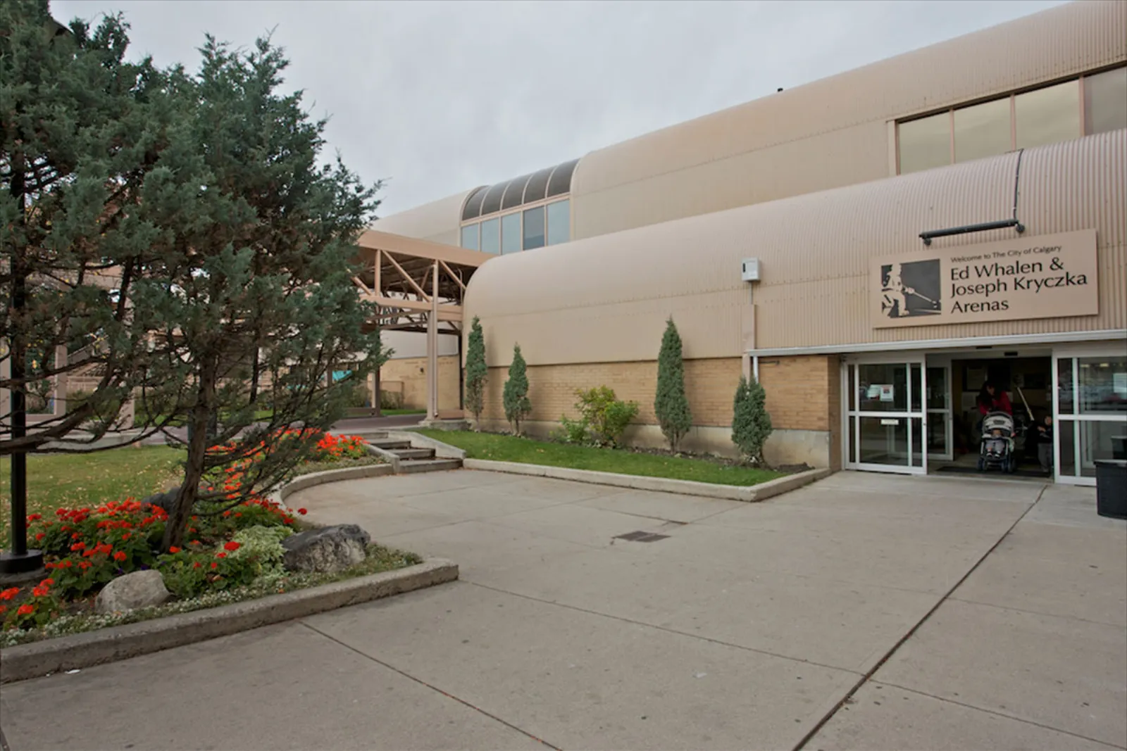 Curved beige metal-clad entrance wing of Southland Leisure Centre with a sign reading "Welcome to The City of Calgary — Ed Whalen & Joseph Kryczka Arenas" beside the doors; landscaped entry walkway in front. — Ed Whalen and Joseph Kryczka Arenas
