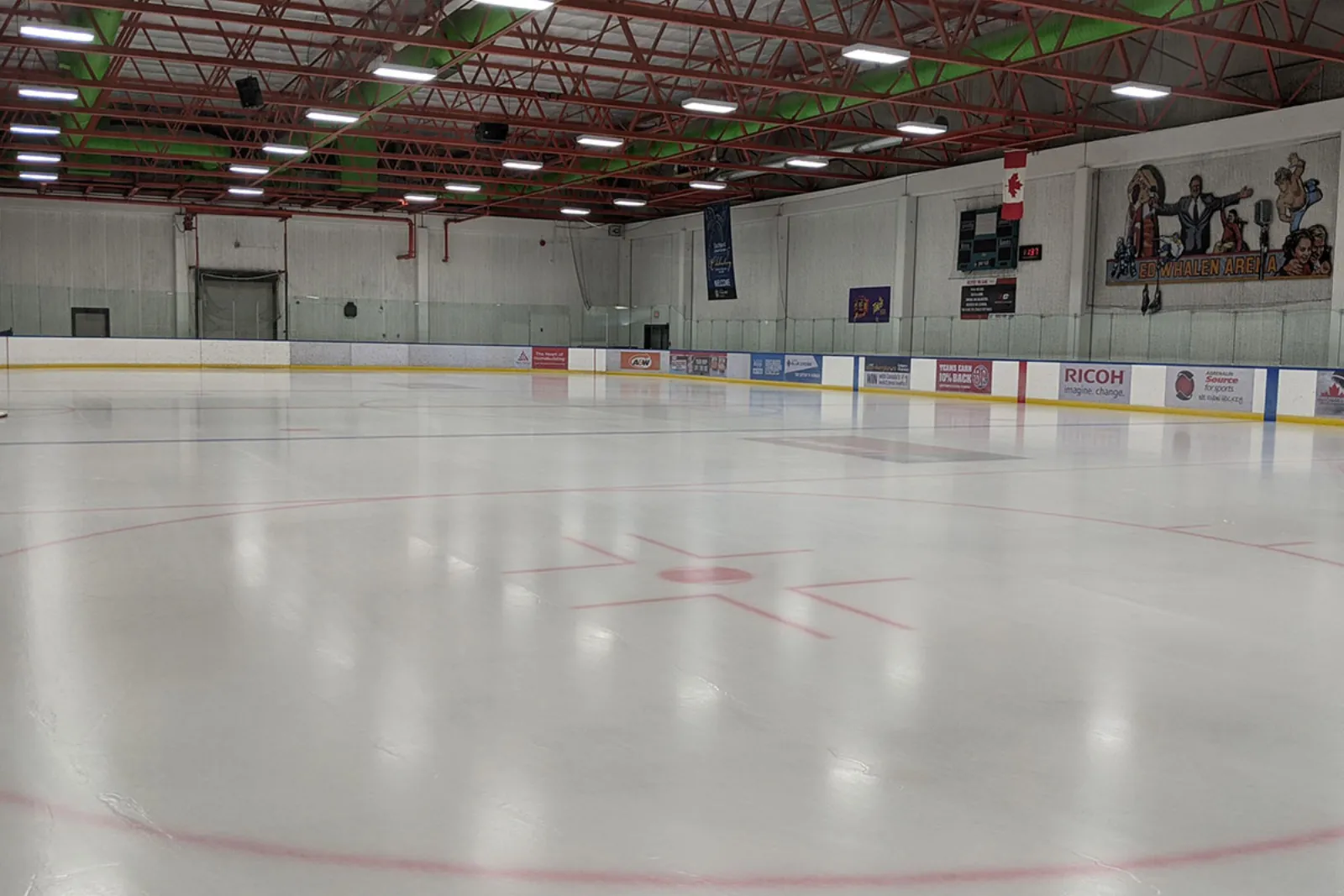 Empty indoor ice rink with red centre-ice circle, rink-board ads and a painted wall mural reading "Ed Whalen Arena", red roof trusses overhead. — Ed Whalen and Joseph Kryczka Arenas
