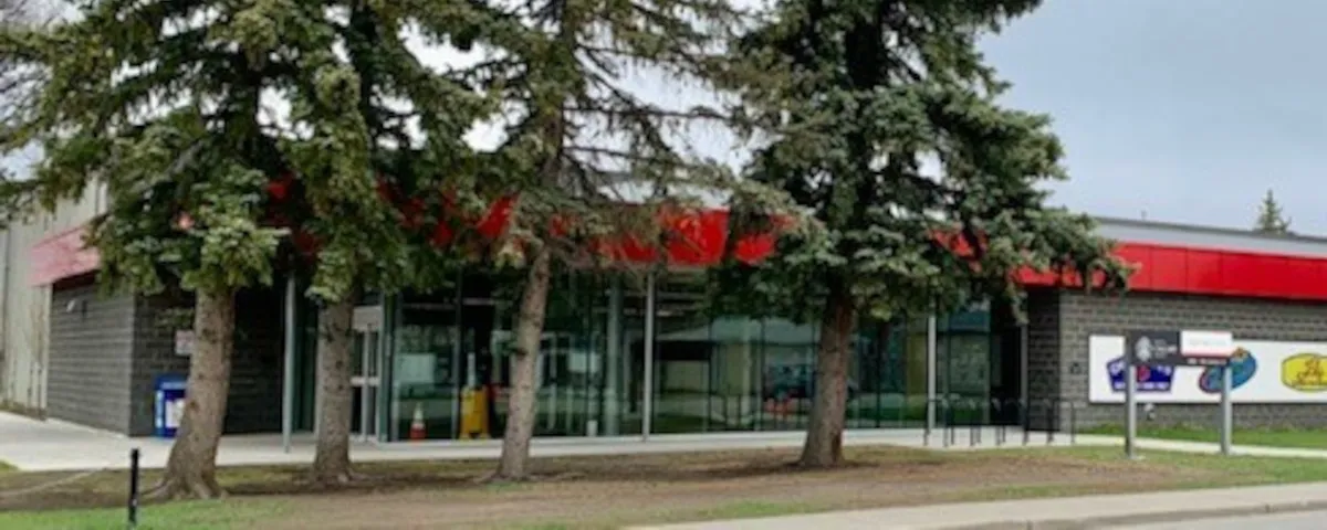 Ernie Starr Arena's glass-fronted entrance with a red fascia band and grey block walls, partly screened by tall spruce trees, with a sponsor sign at right. — Ernie Starr Arena
