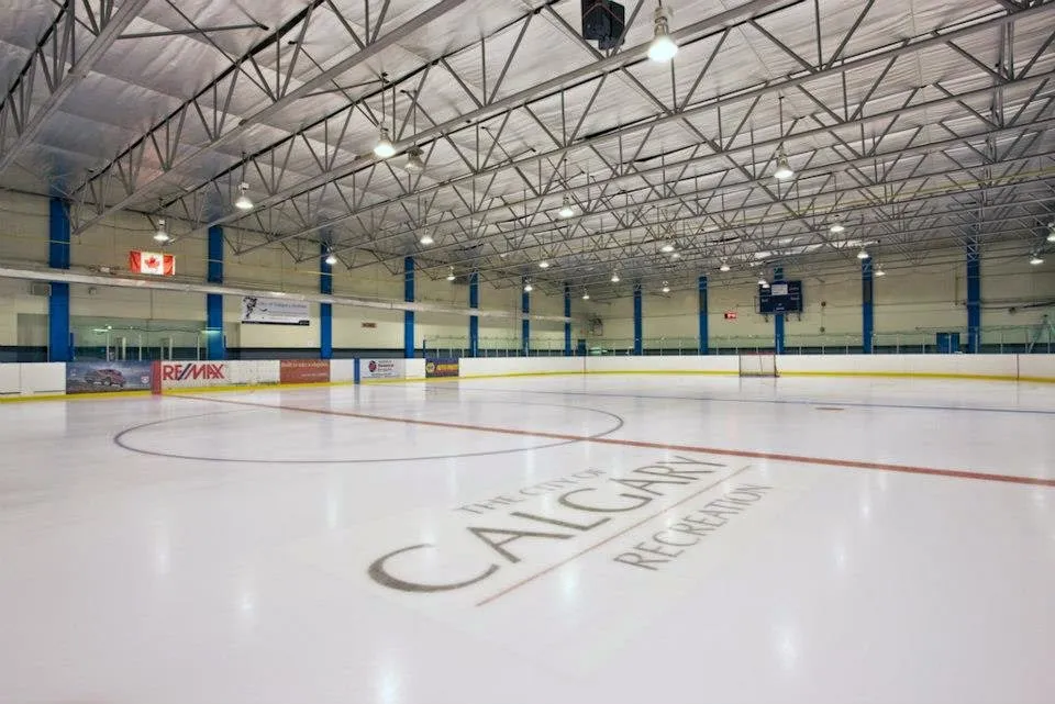 Empty ice at Ernie Starr Arena, City of Calgary Recreation lettered at centre beneath a steel-truss roof and blue columns — Ernie Starr Arena