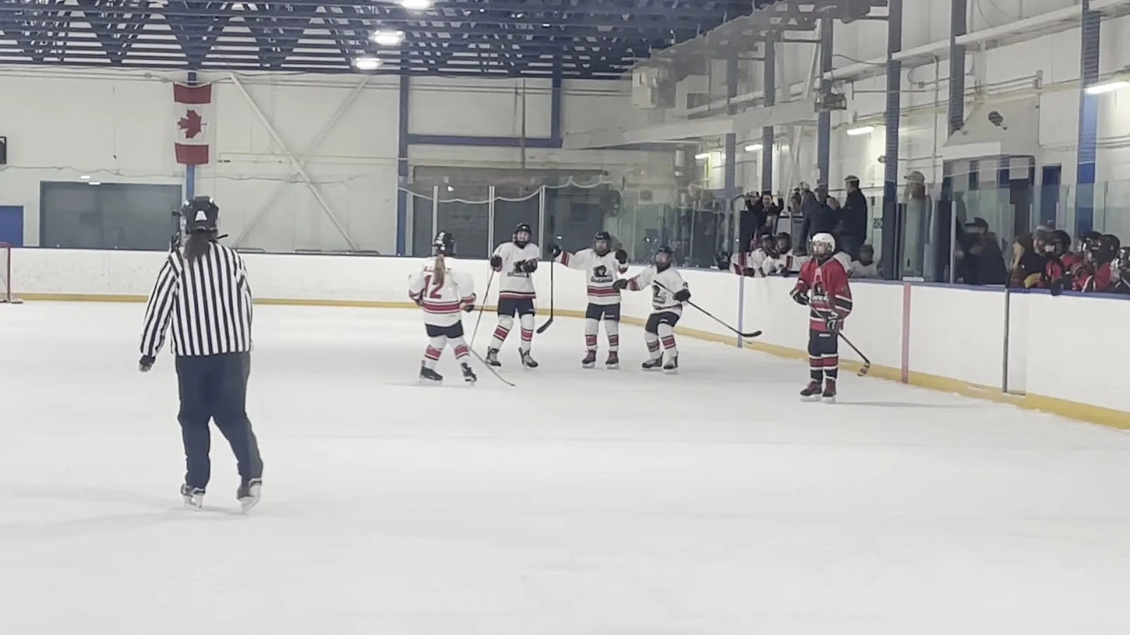 Un partido visto desde el vidrio, con jugadores repartidos por el hielo junto a un árbitro bajo un techo con refuerzos azules y un reloj de marcador al fondo de la pista. — Optimist and George Blundun Arenas