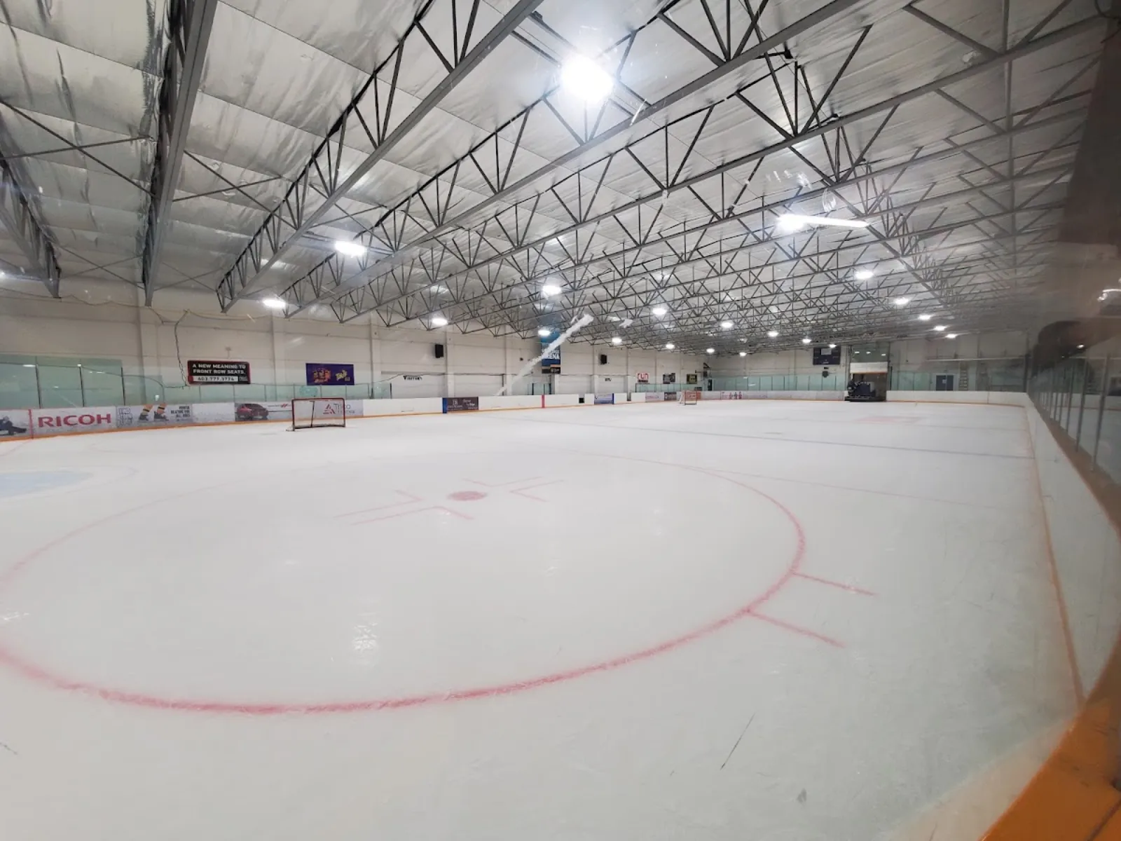 Pista de hielo techada vacía con el círculo rojo del centro de la pista, vallas con cristal y un panel de anuncio de RICOH, bajo armaduras de acero del techo. — Shouldice Arena