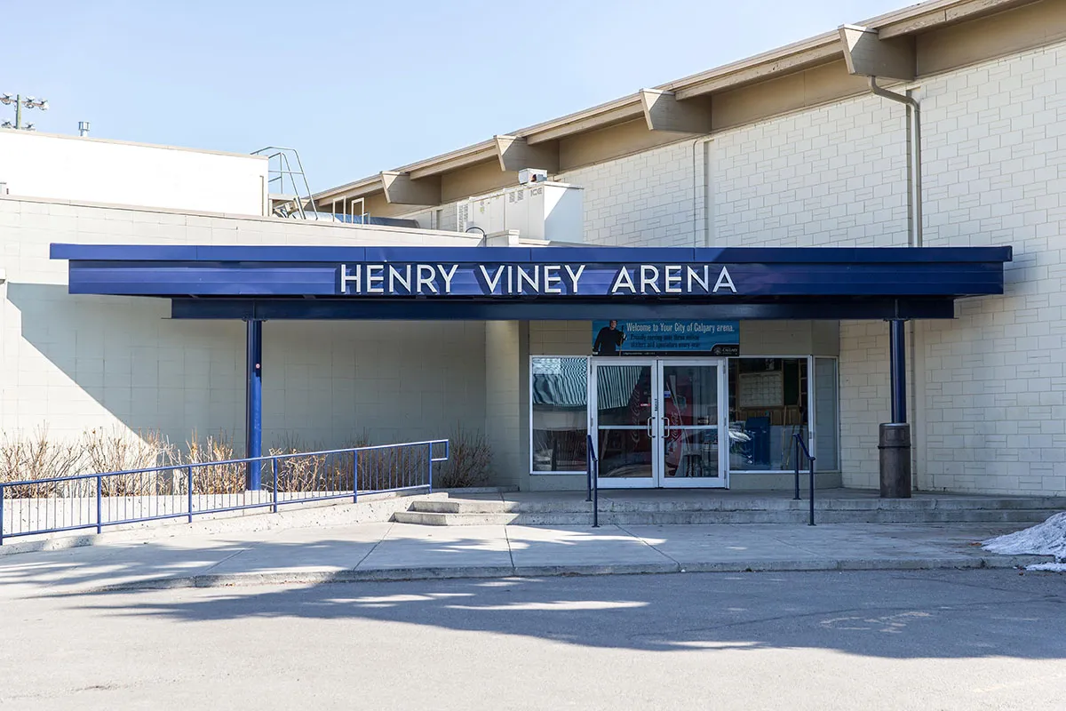 La entrada de Henry Viney Arena: una marquesina azul marino con letras blancas de 'HENRY VINEY ARENA' sobre puertas de vidrio, con escalones y una rampa de barandal azul frente al edificio de bloques claros. — Stew Hendry and Henry Viney Arenas