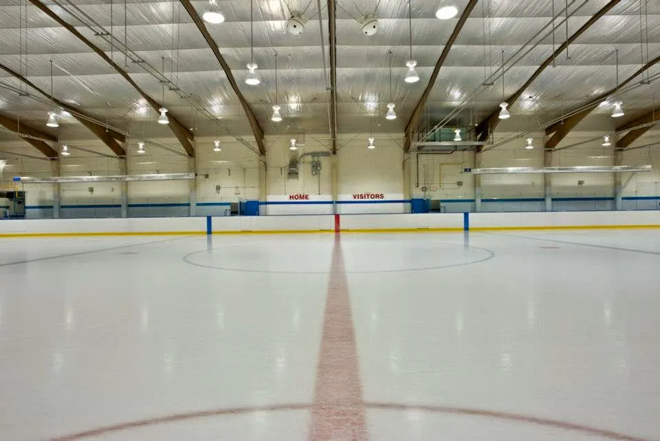 Una pista de hielo vacía dentro de la arena, a lo largo de la línea roja central hacia las vallas del fondo marcadas HOME y VISITORS bajo un techo en arco de armaduras de madera y acero. — Stew Hendry and Henry Viney Arenas