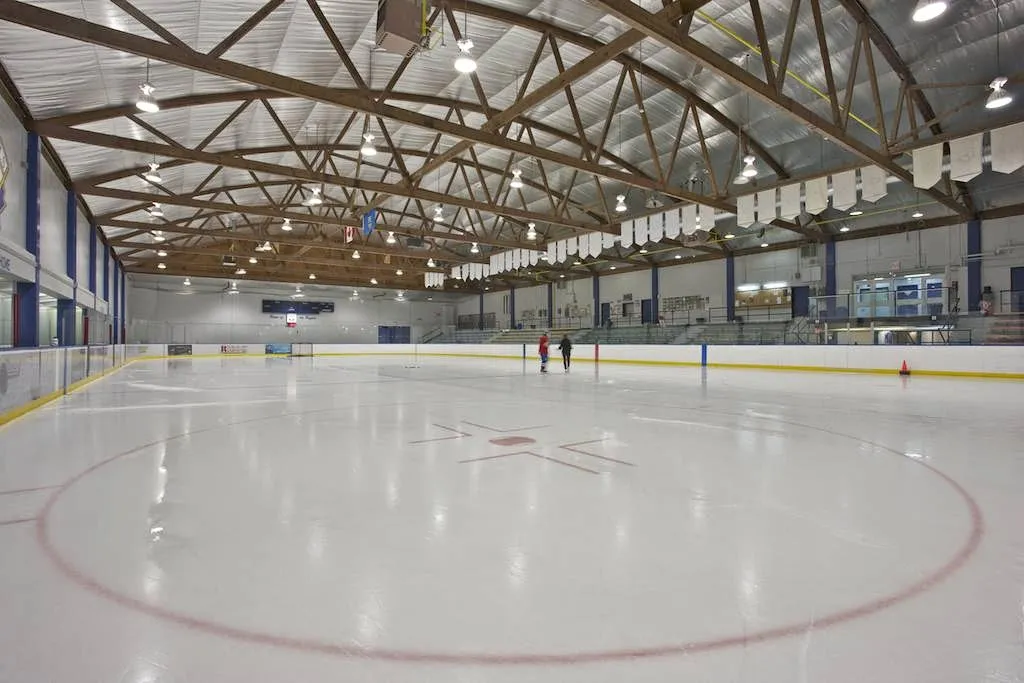 Empty full ice sheet under an exposed wood-truss roof, viewed from the stands with two skaters near center ice and bleacher seating along the far side. — Stu Peppard Arena