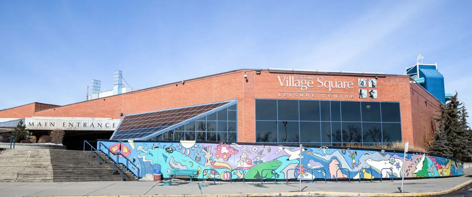 Brick main entrance of Village Square Leisure Centre, the shared building housing the Village Square and Wally Kozak arenas, with a "Village Square Leisure Centre" sign and skating/hockey pictogram icons on the wall above the main entrance. — Village Square and Wally Kozak Arenas