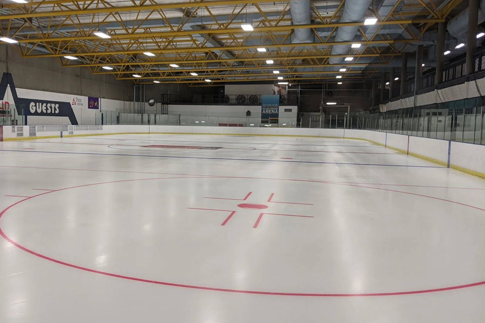 Empty indoor ice rink with a blue banner reading "Welcome to the City of Calgary Village Square Arena", spectator railing above the boards, and yellow roof trusses overhead. — Village Square and Wally Kozak Arenas