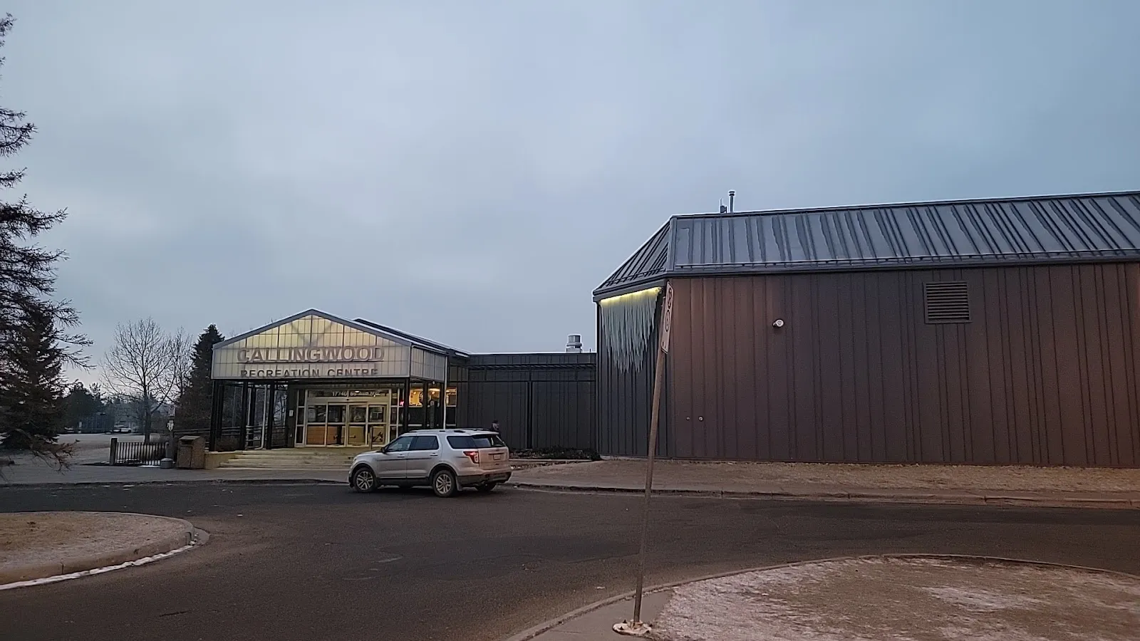 The dark-clad timber side of the Callingwood Twin Arenas with a yellow-fronted entrance block beside a car park under an evening sky. — Callingwood Twin Arena