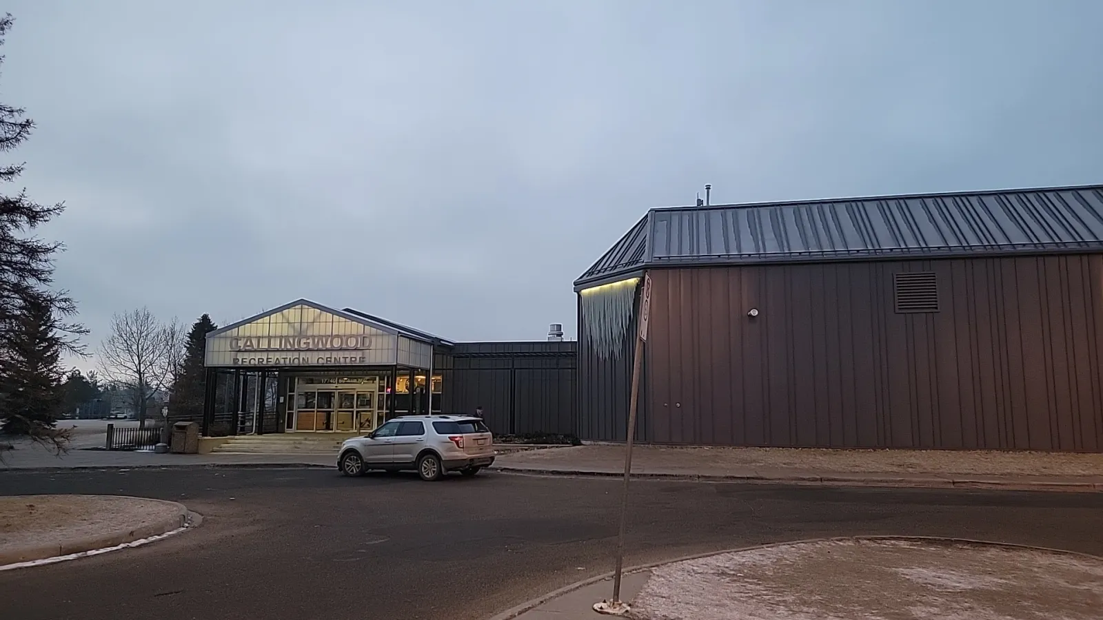 The lit glass entrance pavilion between the two brown metal-clad rink buildings, seen across the parking lot at dusk. — Callingwood Twin Arena